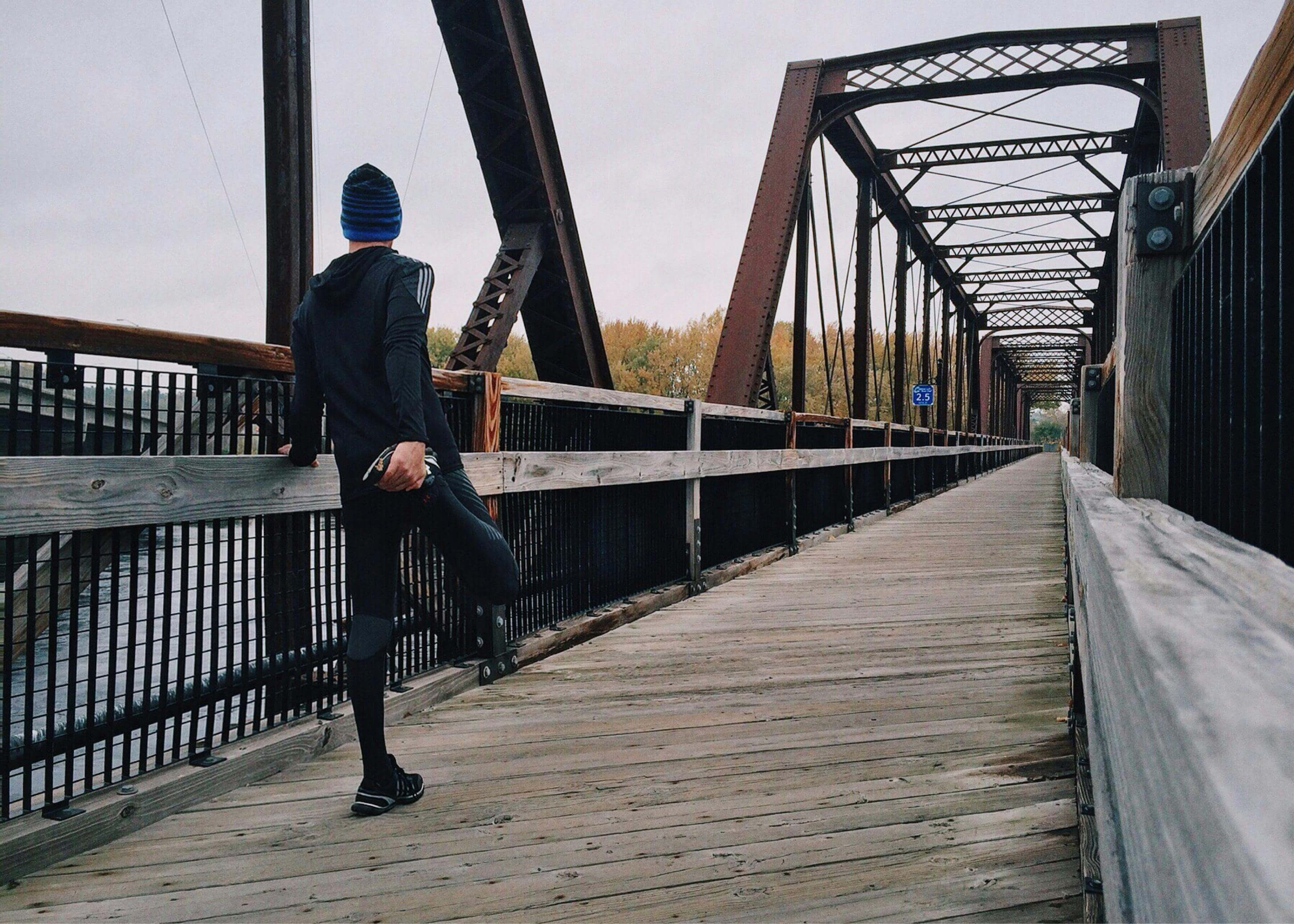 Person standing on a wooden bridge with metal railings and a bridge in the background.