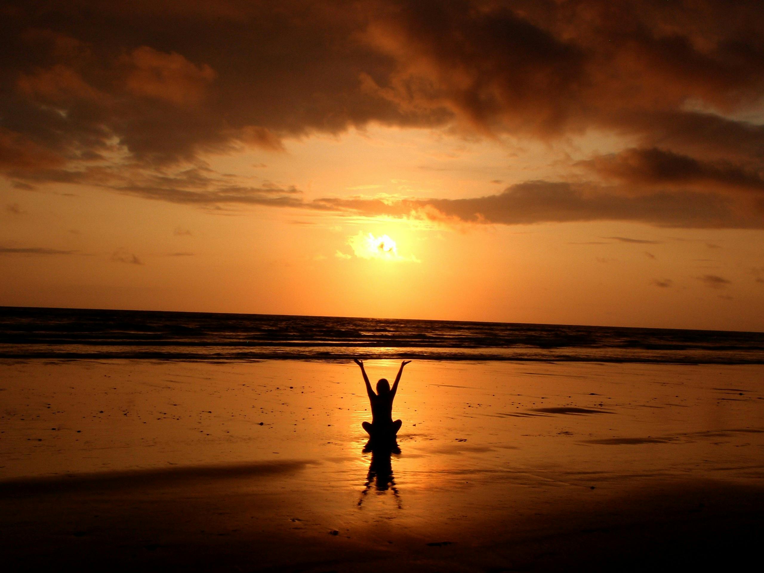 Person sitting on a beach at sunset with arms raised, silhouetted against the orange sky.