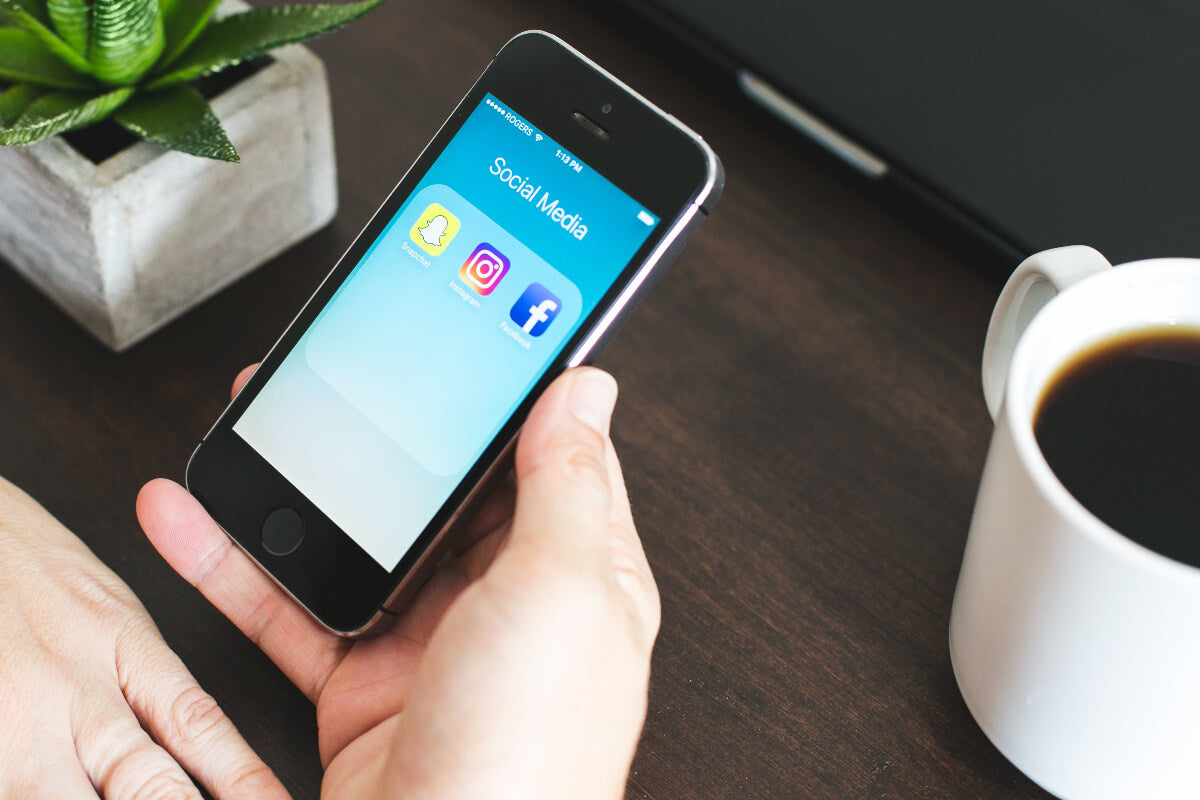 Person holding a smartphone with social media app icons on a desk with a cup of coffee and plant.